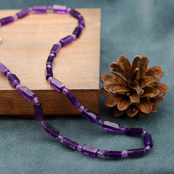 Purple beaded necklace on a wooden block with a pine cone on a green surface
