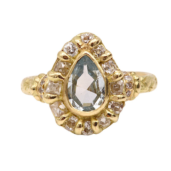 A yellow gold ring featuring a teardrop-shaped light blue gemstone surrounded by a halo of small clear stones, set against a plain white background.