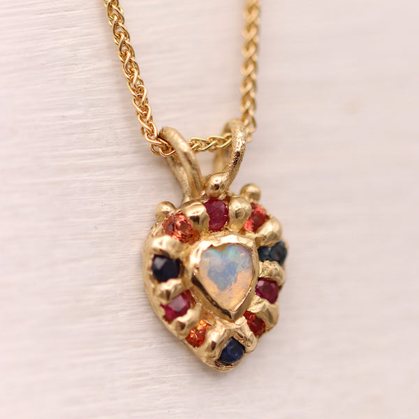 A gold heart-shaped pendant necklace featuring a central iridescent opal heart and surrounded by small red and dark blue gemstones, displayed on a light textured background.