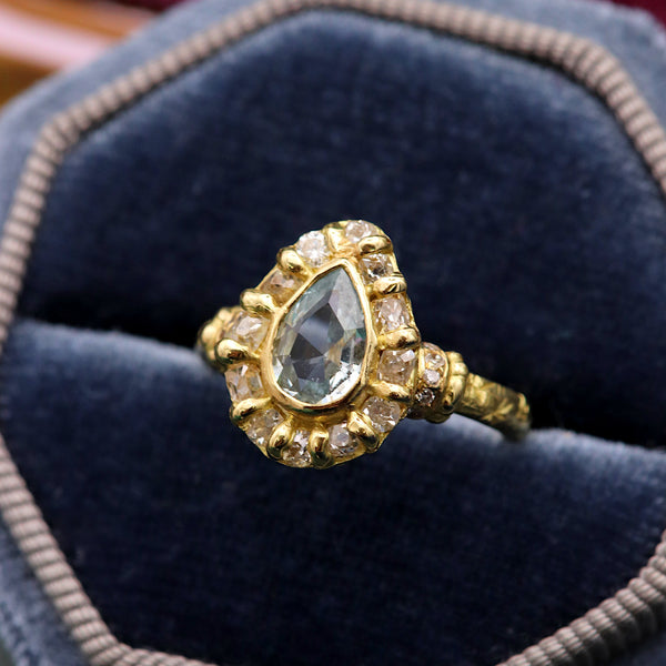 A gold ring with a teardrop-shaped blue gemstone and surrounding small diamonds, displayed in a blue velvet ring box.