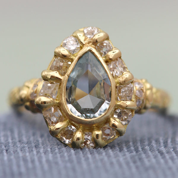 A vintage-style yellow gold ring featuring a pear-cut gray gemstone in the center, surrounded by a halo of small clear stones, displayed on a textured gray surface.