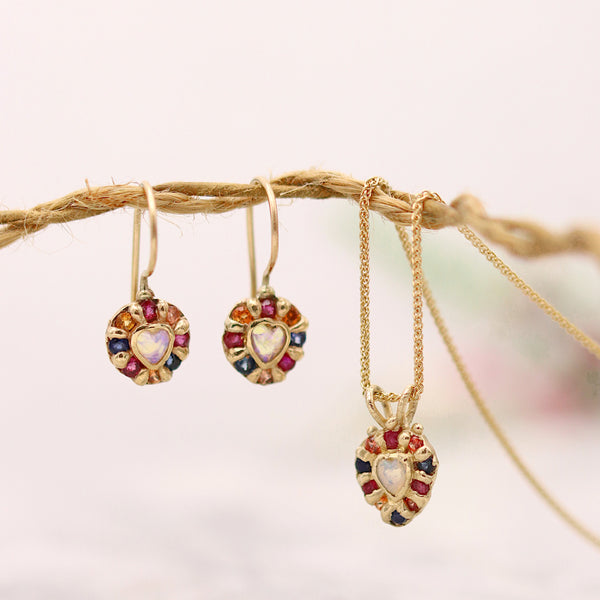 Gold jewelry set featuring a matching pendant necklace and earring pair, each adorned with a central heart-shaped stone and surrounded by red, blue, and yellow gemstone accents, hanging from a rustic twine branch against a soft, light background.