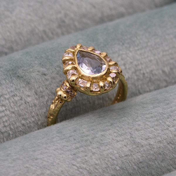 A vintage-style gold ring with a pear-cut light blue center stone and a halo of small grey stones, displayed on a soft gray velvet surface.