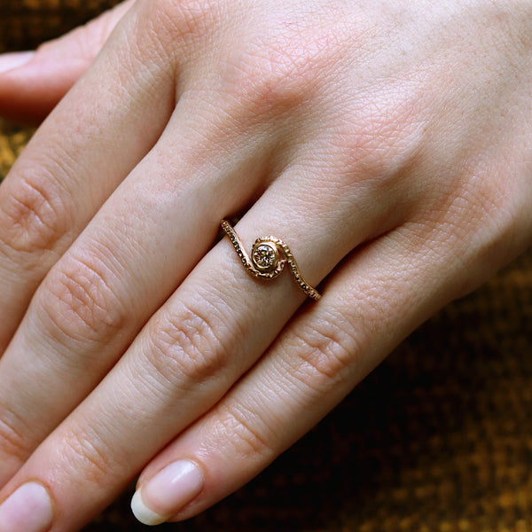 A close-up of a hand wearing a delicate gold ring with a small rose-shaped center and tiny clear stones, set against a dark textured background.