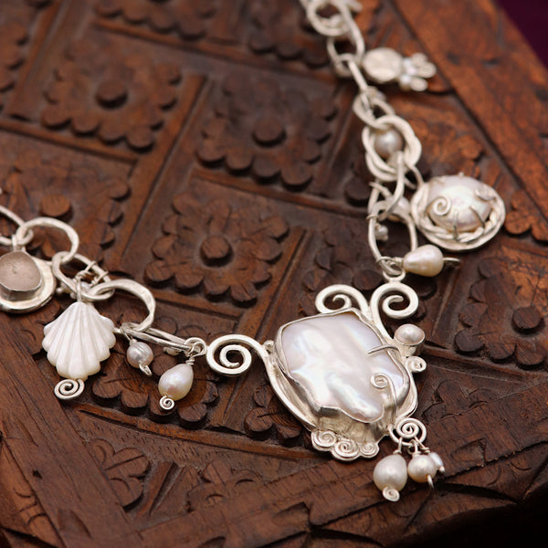 A silver-toned shell-themed necklace with mother-of-pearl shell charms and small pearl beads, displayed on a carved dark brown wooden box.