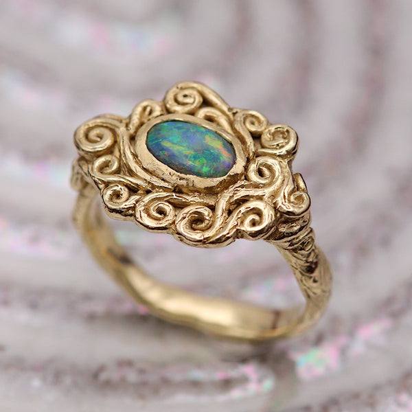 A gold ring with an ornate swirling design and a central oval opal gemstone showing blue and green iridescence, resting on a textured, iridescent surface.
