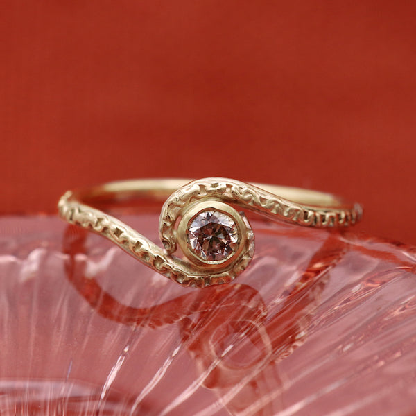 A gold ring with a textured, swirling band and a central round rose-gold gemstone, resting on a textured pink surface against a solid red background.