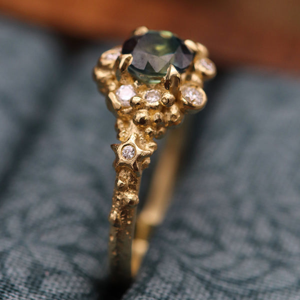 A close-up of a gold ring with a dark green round center stone and small clear accent stones, resting on a textured blue-gray fabric surface.
