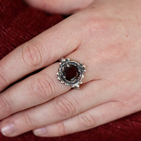 Hand wearing a silver ring with a red stone on a red fabric background
