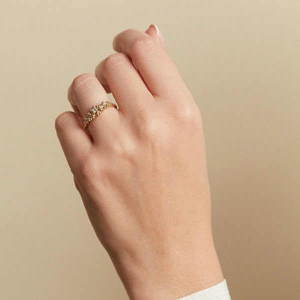 A close-up of a hand wearing a delicate gold ring with small clear stones, set against a soft beige background.