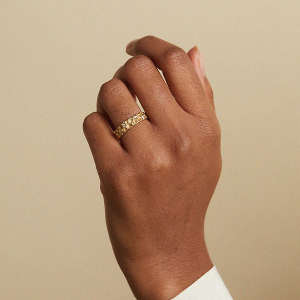 A close-up of a person's hand wearing a delicate gold band ring with small clear stones, set against a plain beige background.