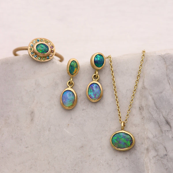 A gold jewelry set featuring a ring, drop earrings, and pendant necklace with iridescent green-blue opal-like stones, arranged on a light stone surface.