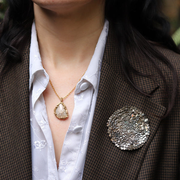 Person wearing a brown blazer with a white shirt, gold necklace, and silver brooch.